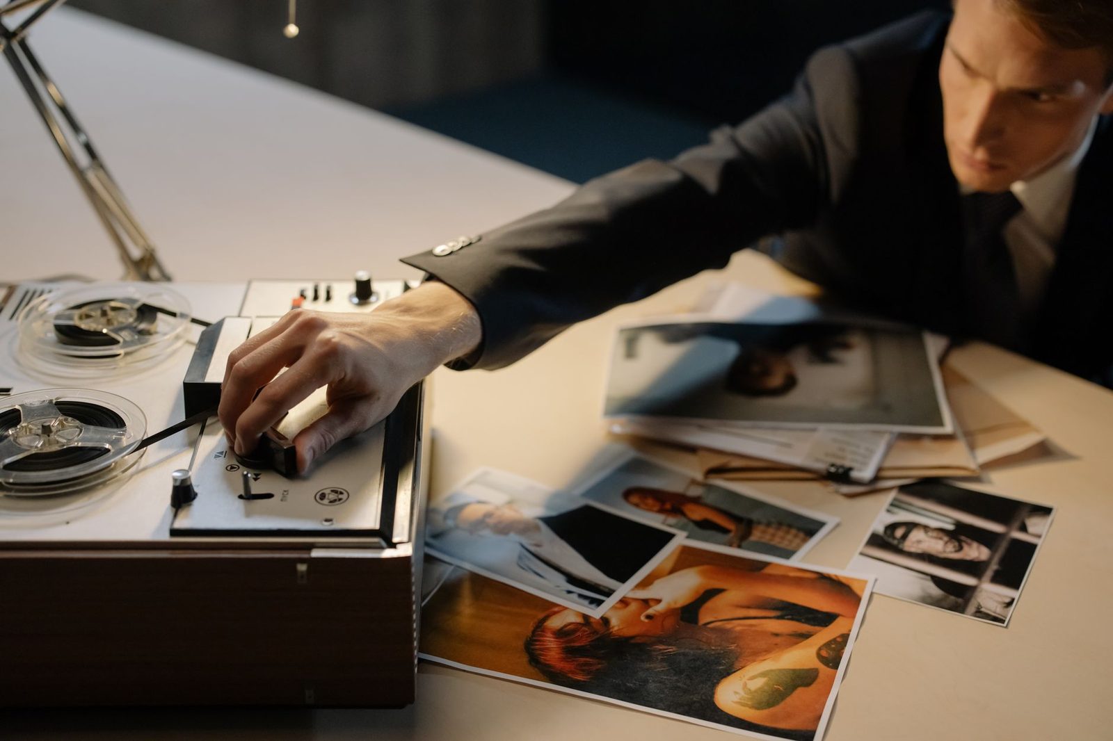 A detective adjusts a tape recorder while photos are spread across a table during an investigation.