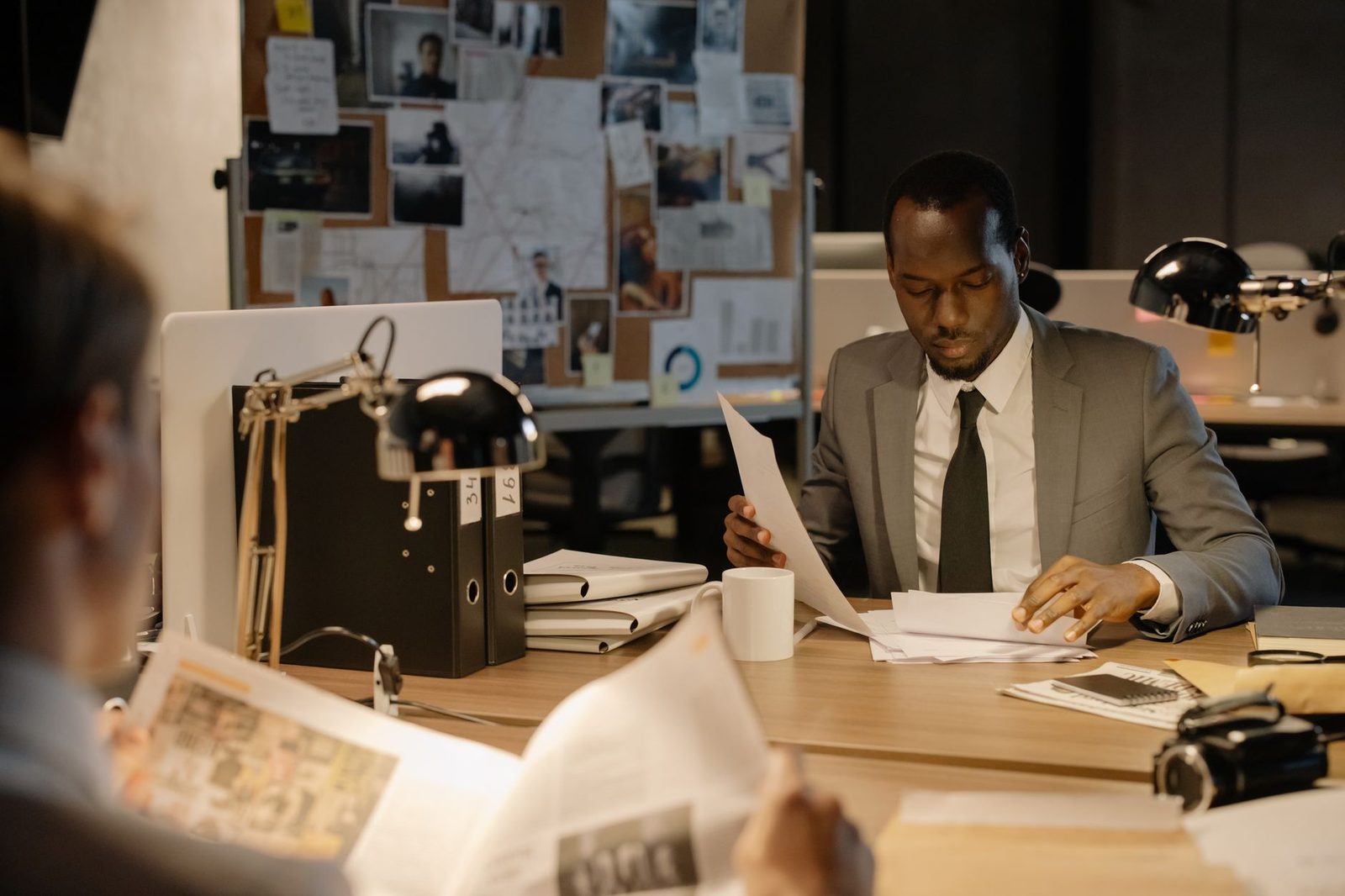 A detective in a suit reviews papers at a desk in a dimly lit office.