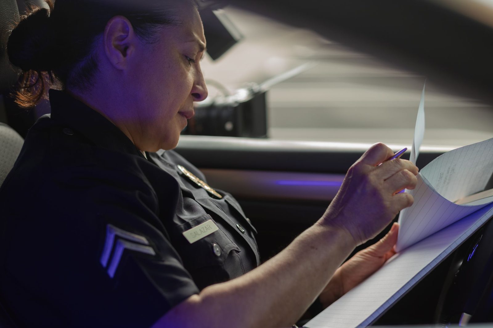 A female police officer sits in a patrol car in daylight, writing a report.