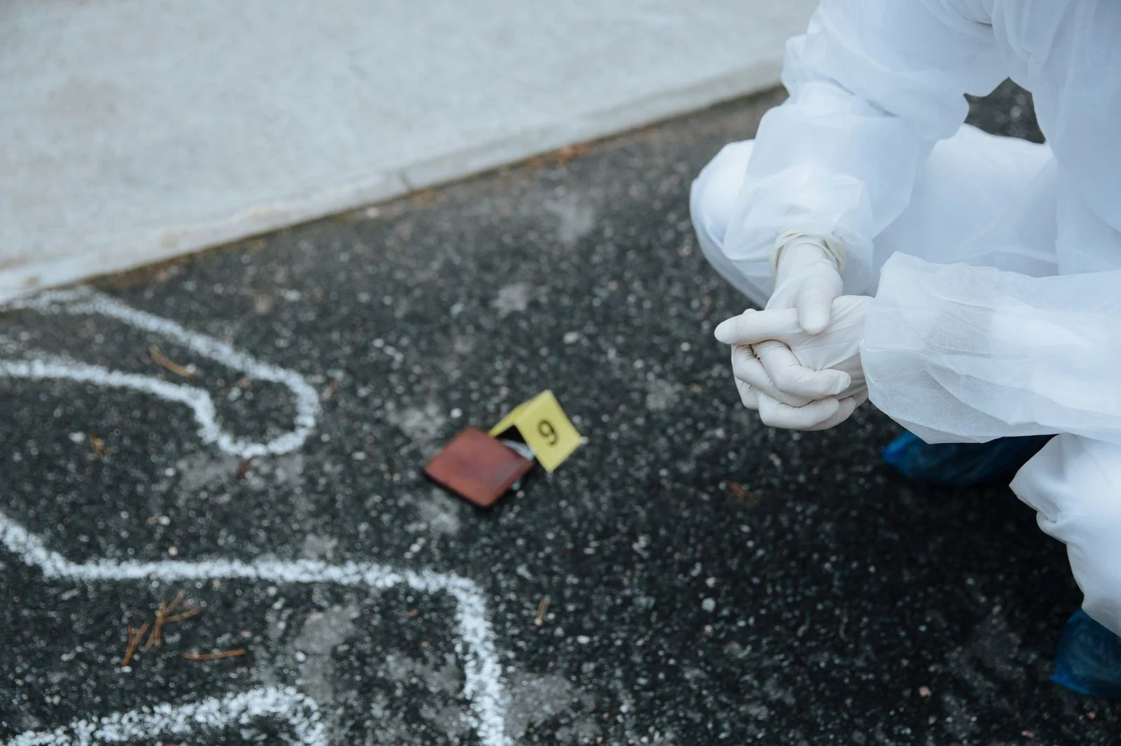 Un investigador forense con traje protector blanco examina el contorno marcado de una escena del crimen sobre el asfalto.