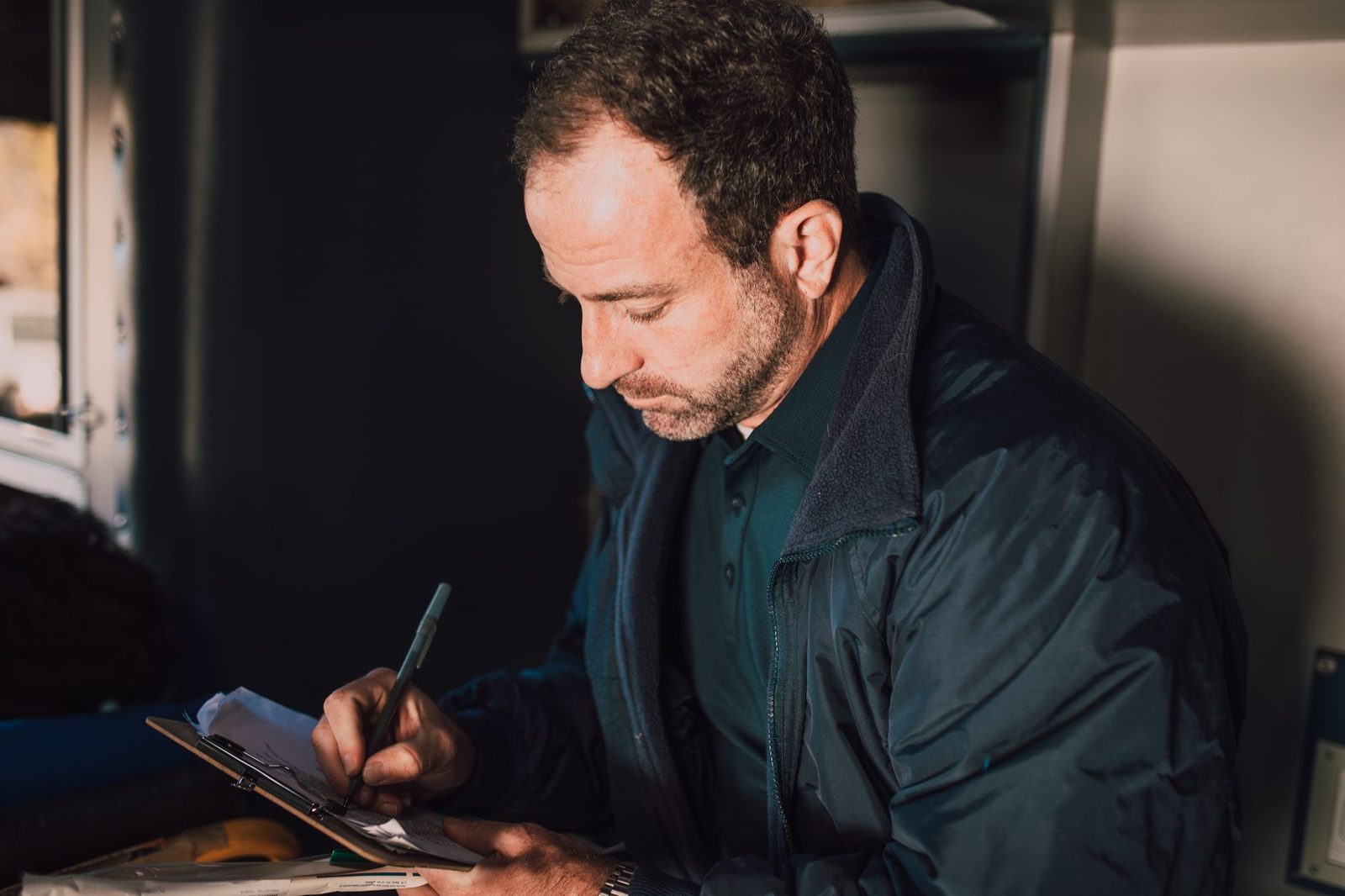 A man in a room writes notes on a clipboard, looking focused and thoughtful.