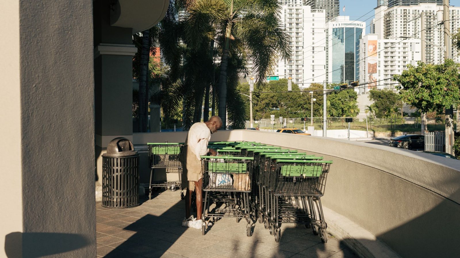 A man pushes shopping carts past high-rise buildings under bright sunlight in a city street.