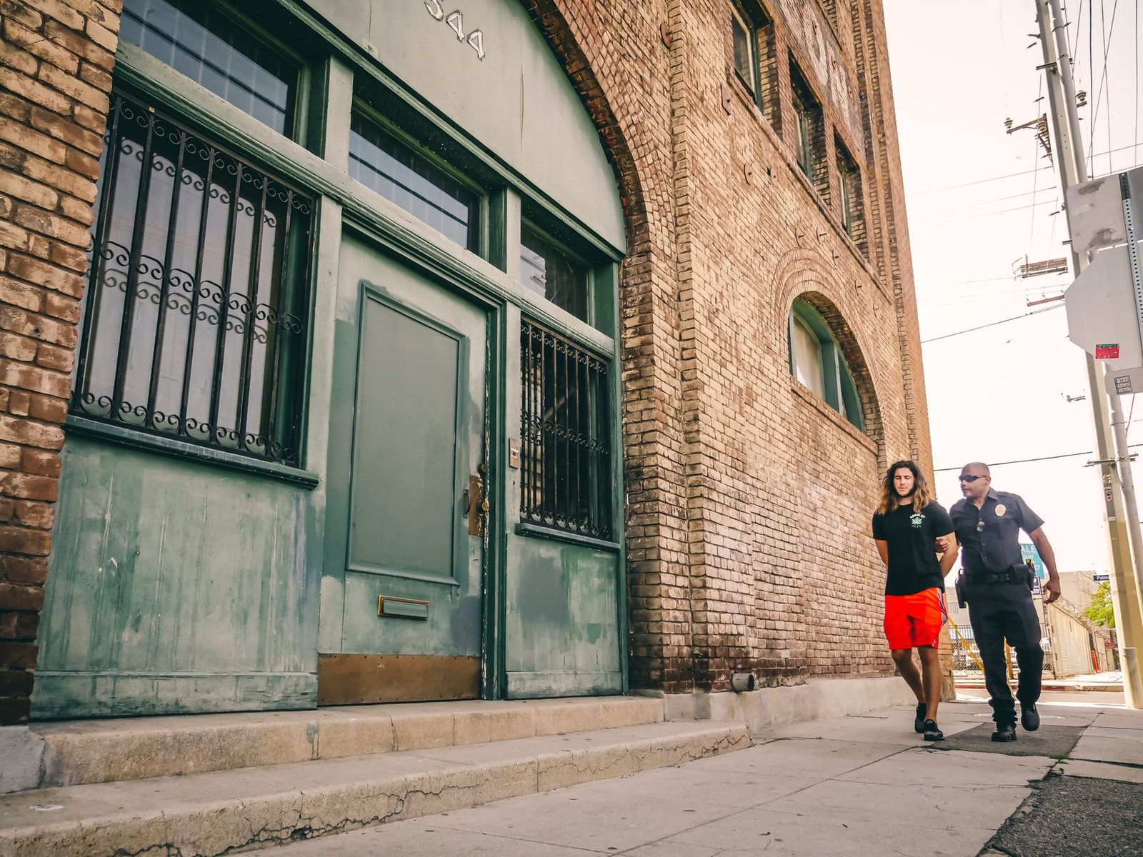 A police officer leads a handcuffed person past a red brick building.