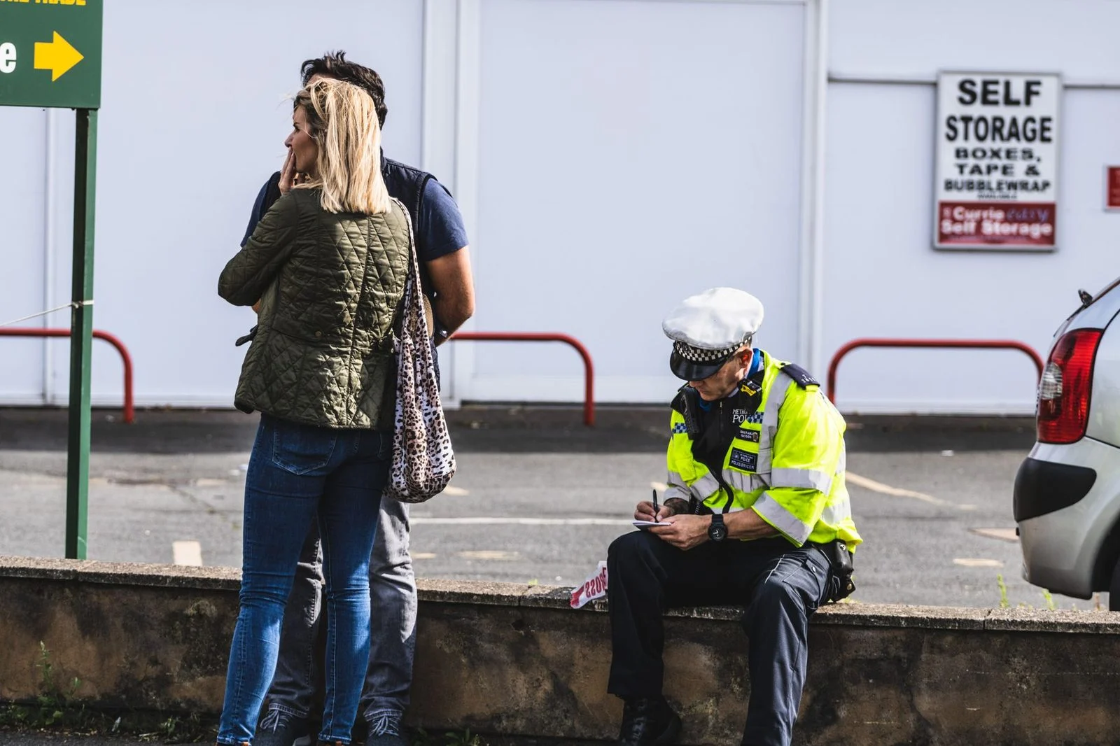 Un agente de policía escribe una multa cerca de un área de almacenamiento, con personas al fondo.