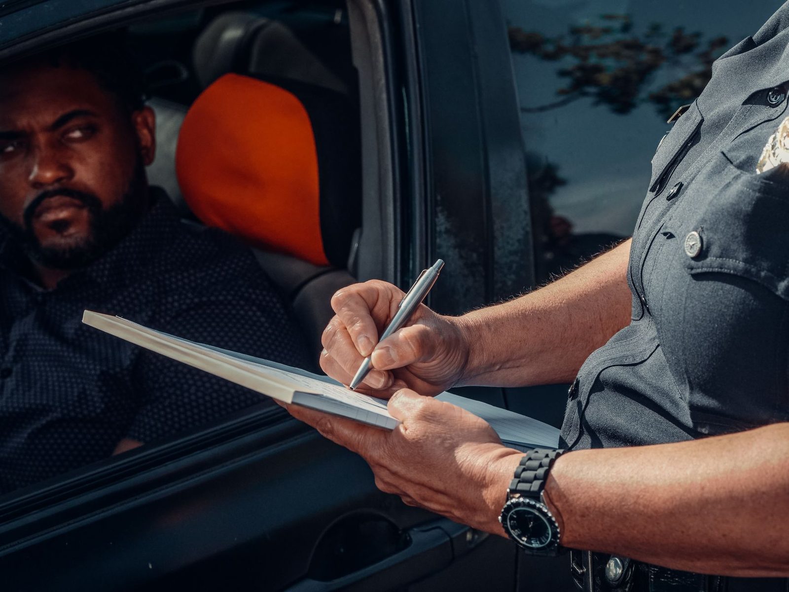 A police officer writes a traffic ticket while a driver watches from inside a parked car.