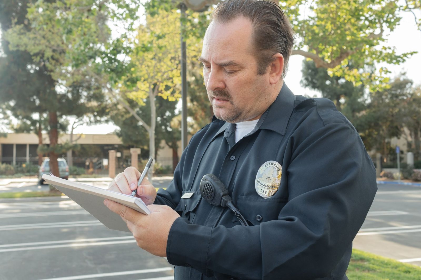 Un agente de policía toma notas en una libreta al aire libre, con árboles al fondo.