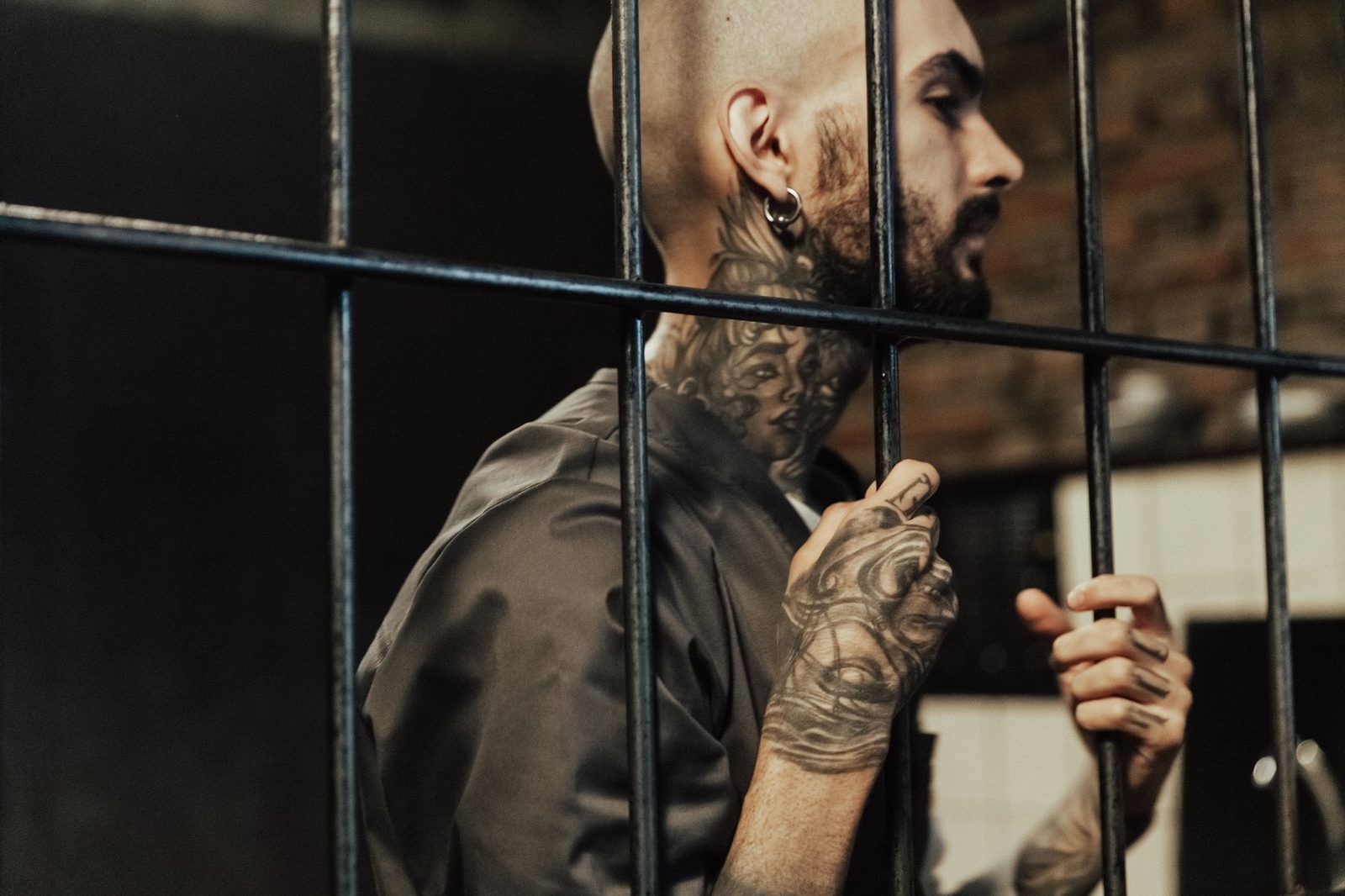 Hombre con tatuajes de pie tras barrotes de una celda, mirando pensativo a lo lejos.