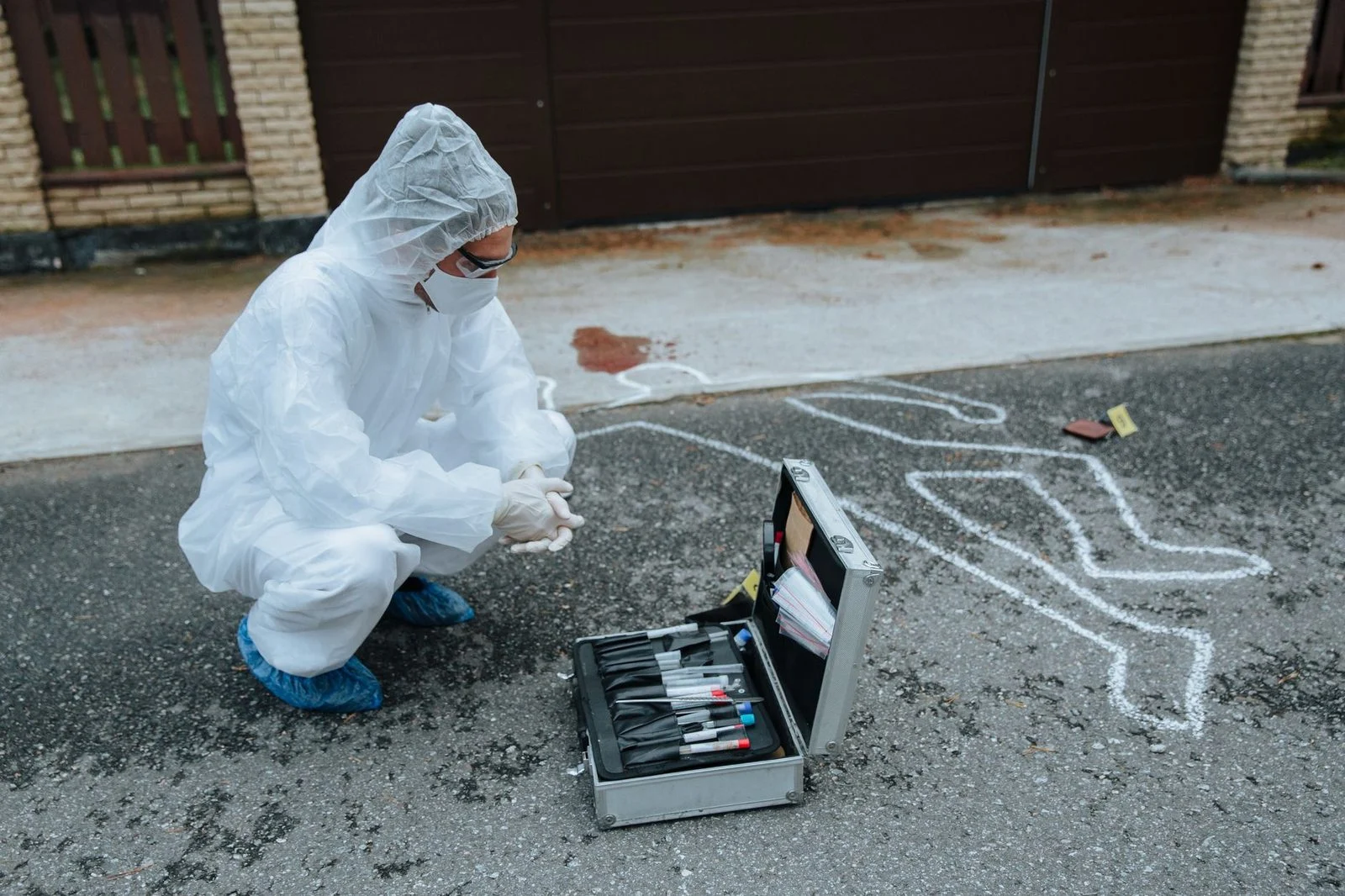 Crime scene investigator in protective gear examining an outdoor scene beside an evidence case.