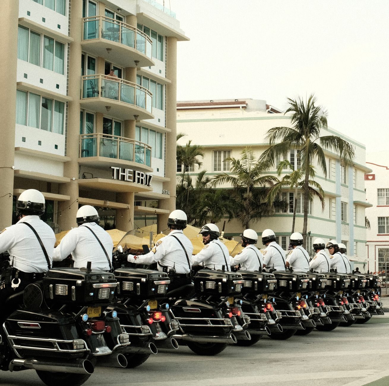 Motorcycle police officers ride in a line through Miami Beach, with urban buildings and palm trees behind them.