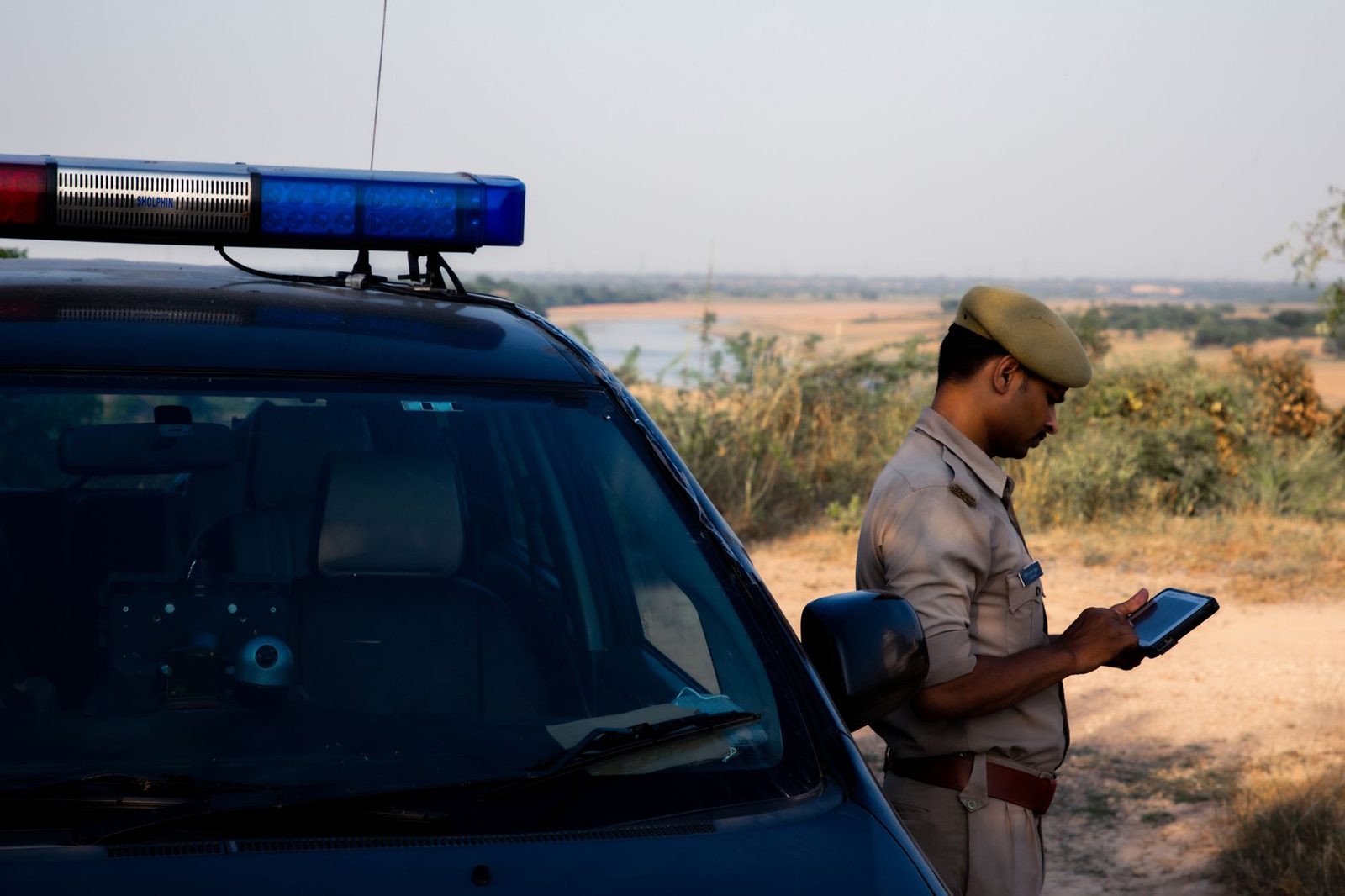 A police officer uses a tablet beside a patrol car on a rural road.