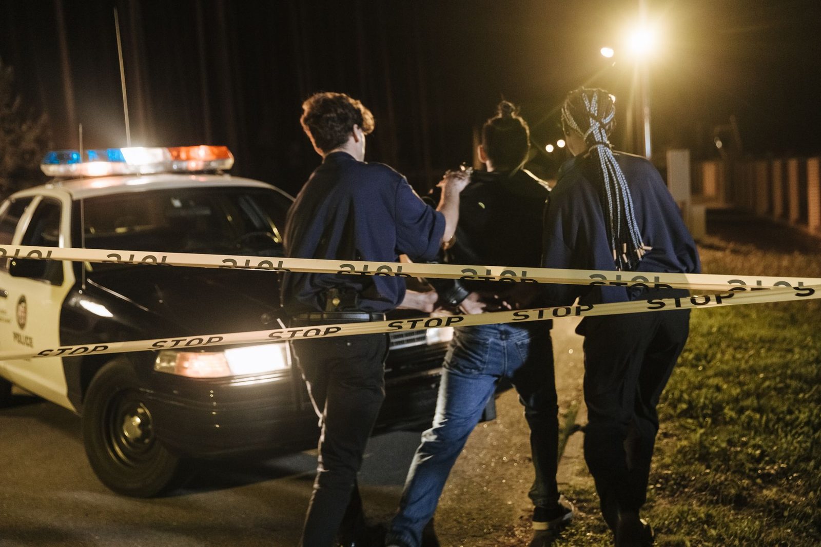 Police officers and a patrol car stand behind caution tape at a nighttime arrest scene.