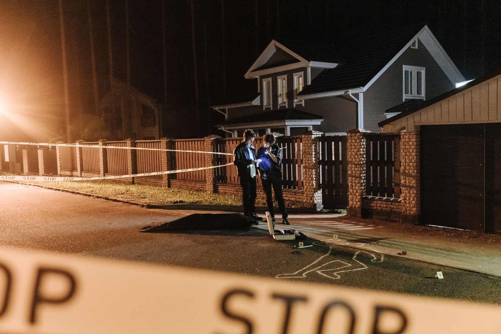 Police officers work a nighttime crime scene outside a house, with barrier tape and evidence markers on the lawn.