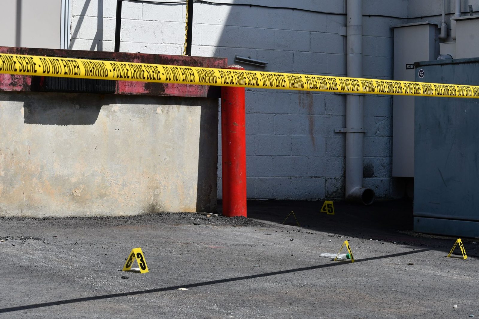 Sunlit city street with yellow caution tape and numbered evidence markers at a crime scene.
