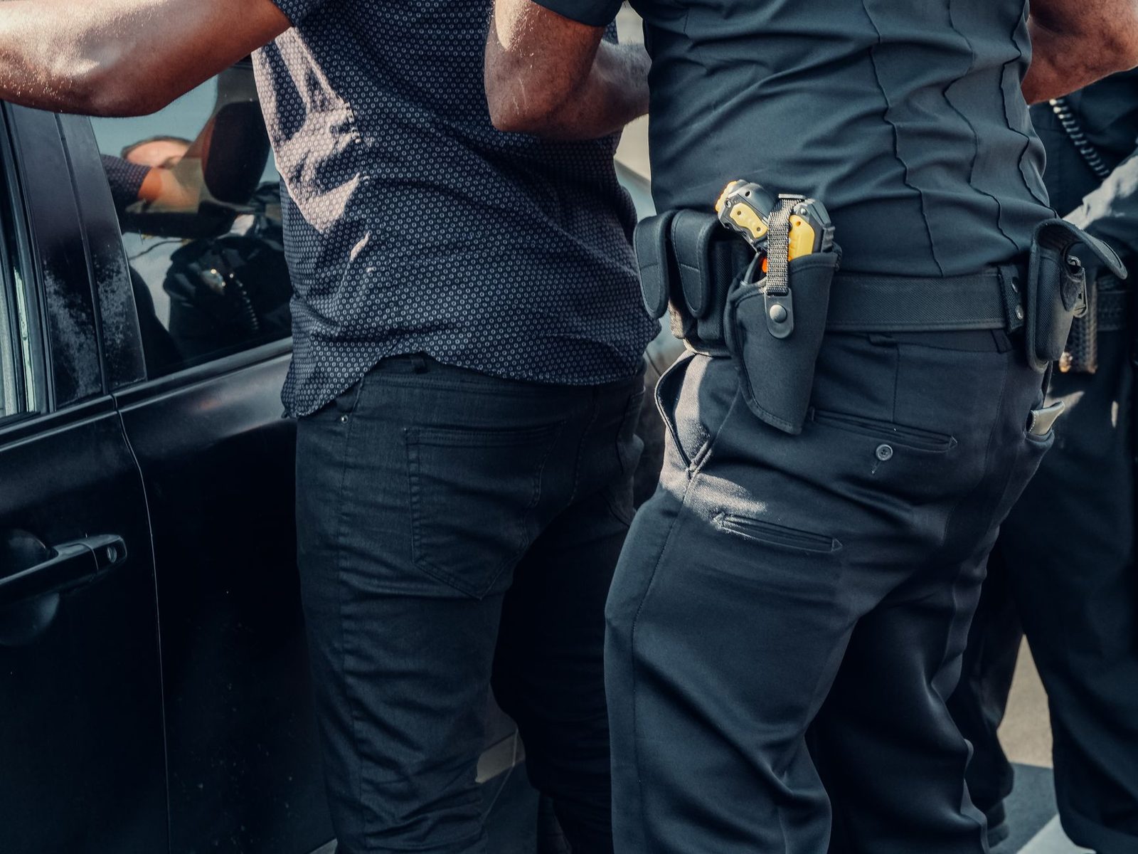 Two police officers detain a person beside a parked car in daylight.
